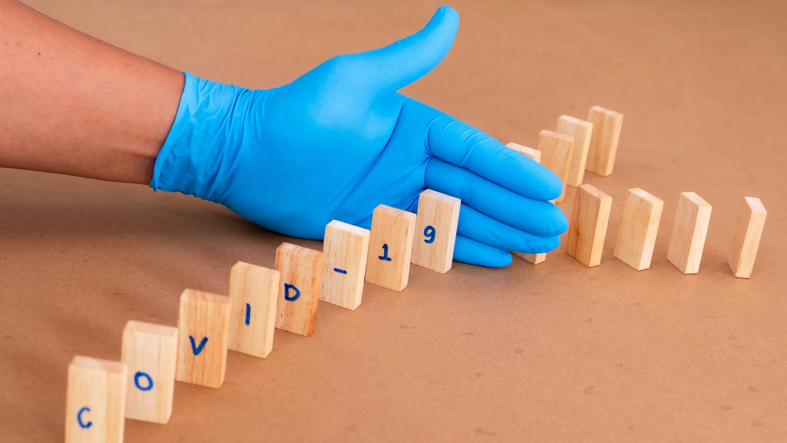 Dominos spelling word Covid-19. A gloved hand protects other dominos from the COVID labeled dominos.