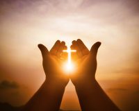 Picture of two hands outstretched, cupped with palms facing upward, towards sunrise. Image of sunlit cross formed in cupped hands.