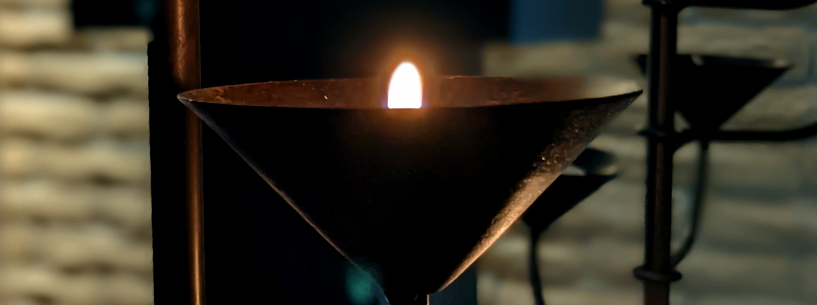 Picture of lit candle in an inverted cone-shaped metal holder. Room is dark.